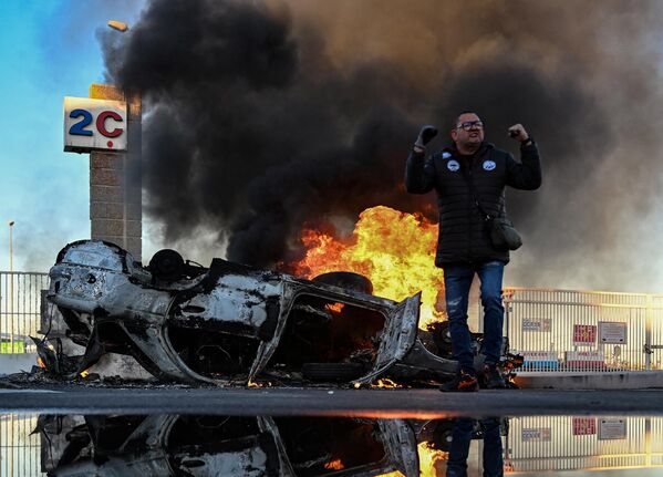 Член профсоюза Всеобщей конфедерации труда стоит рядом с перевернутой горящей машиной во время блокады марсельского порта во Франции - Sputnik Грузия