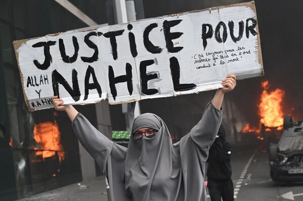 Протестующая держит плакат с надписью &quot;Справедливость для Нахеля&quot; на фоне горящих автомобилей в конце марша памяти в честь подростка, застреленного полицейским в парижском пригороде Нантер. - Sputnik Грузия