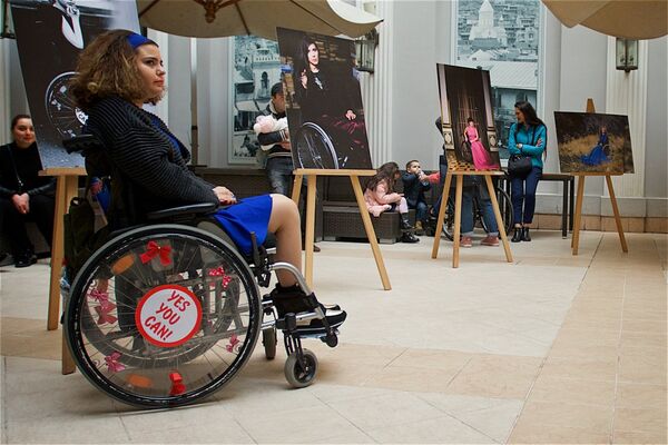 Девушка в инвалидном кресле рядом со своей фотографией на выставке, посвященной Международному женскому дню. - Sputnik Грузия